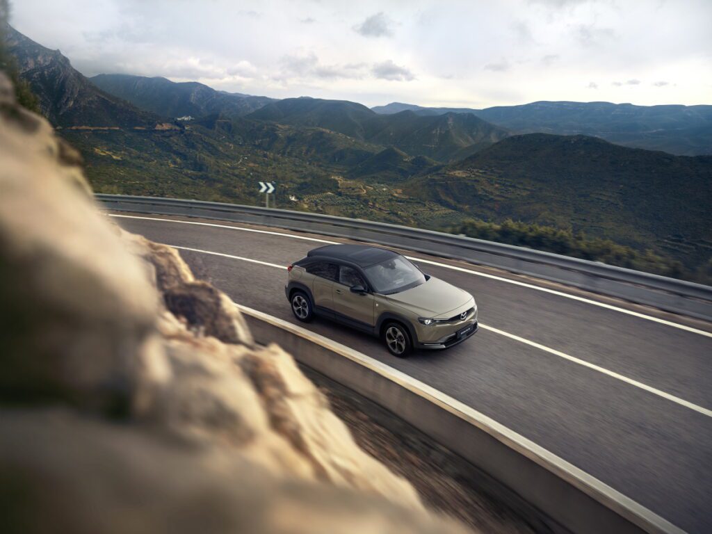 Beżowa Mazda MX 30 z czarnym dachem podczas przejazdu górską drogą, w tle widać doliny oraz wzniesienia